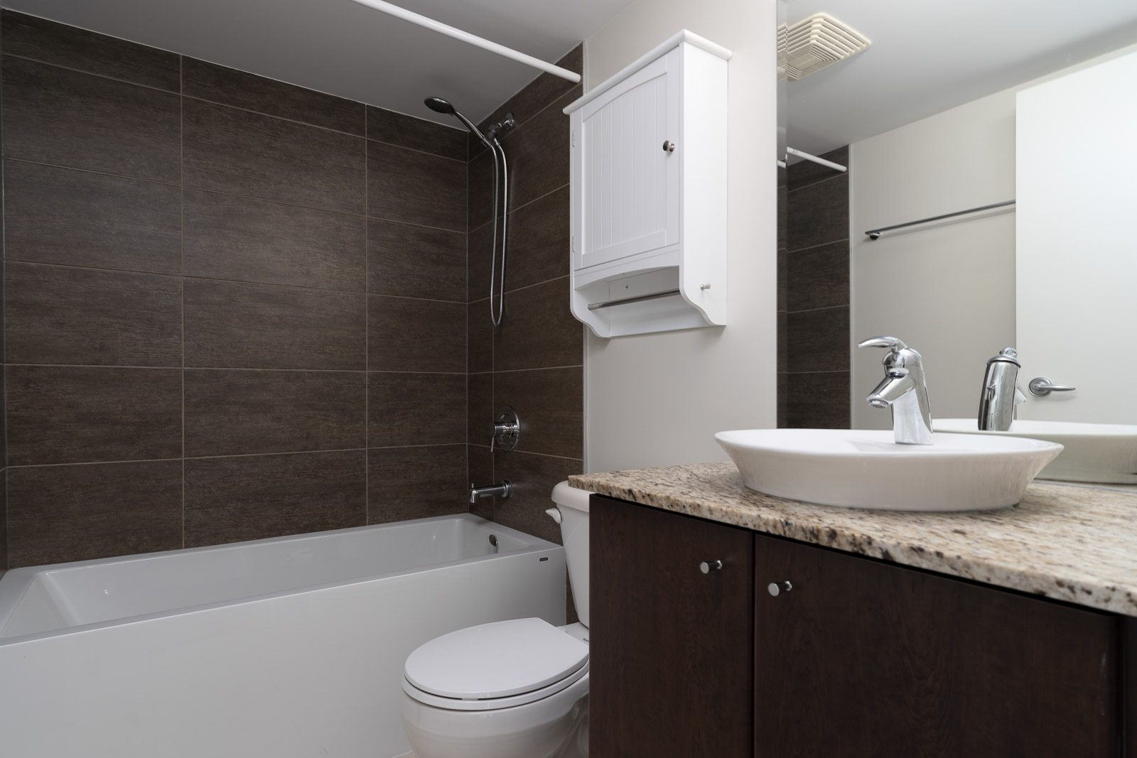 Modern bathroom with a bathtub-shower combo, wall-mounted cabinet, toilet, and sink with a granite countertop and dark wood cabinets.