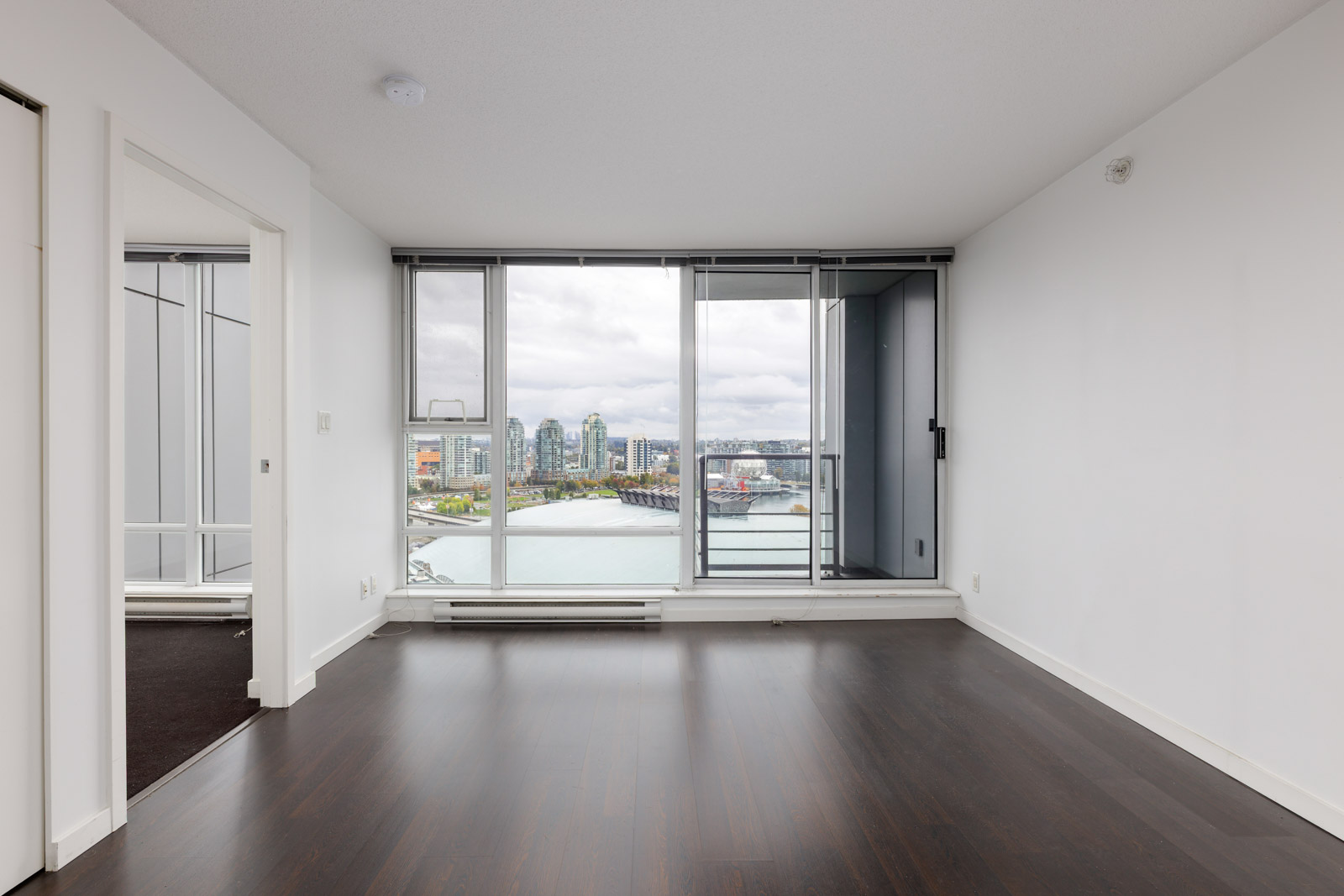 Unfurnished apartment room with dark wood floors, white walls, large windows, and a balcony overlooking a cityscape and waterfront.