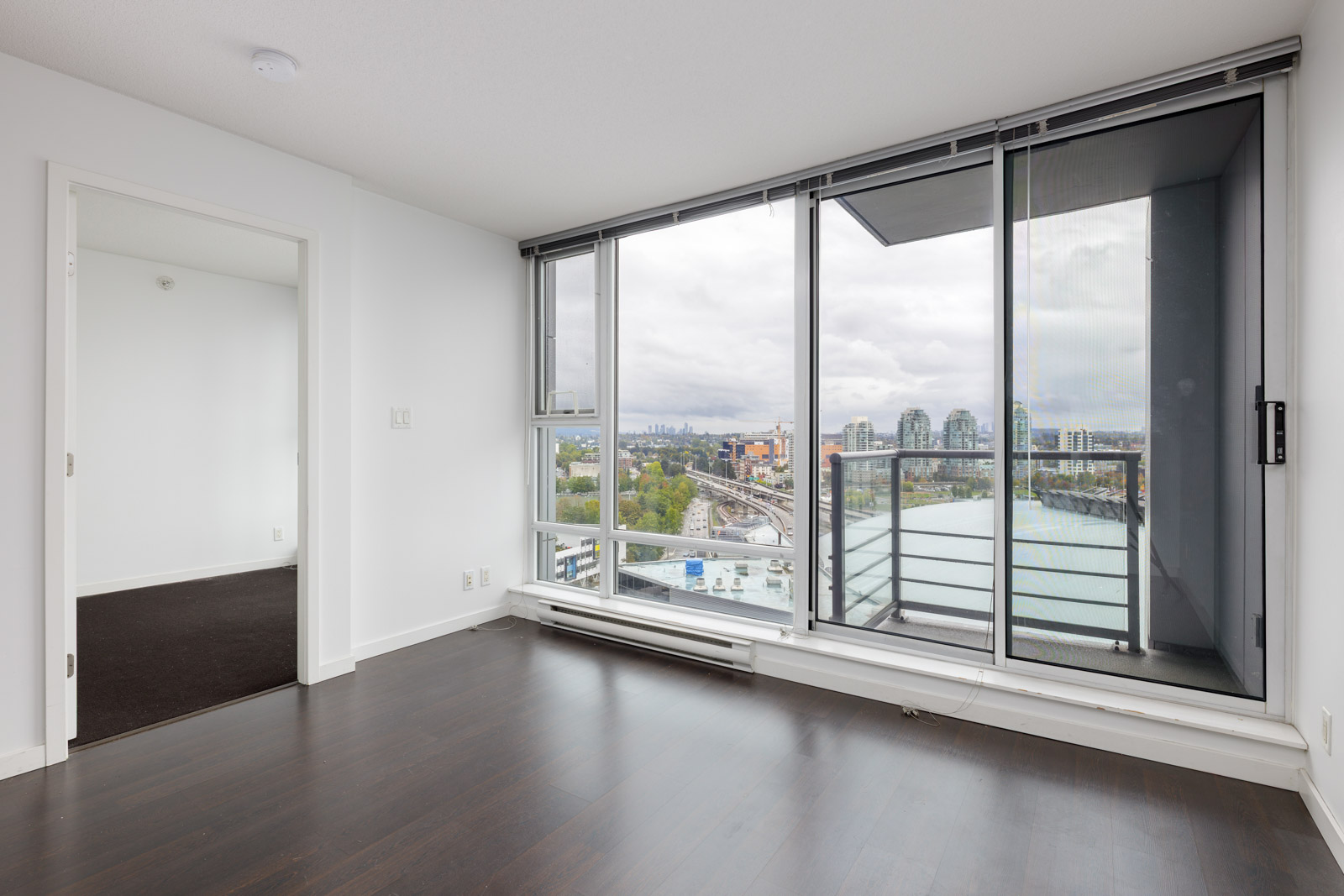 Unfurnished modern apartment with dark wood floors, large windows, balcony, and city and river views. Adjacent room with carpet visible through open door.