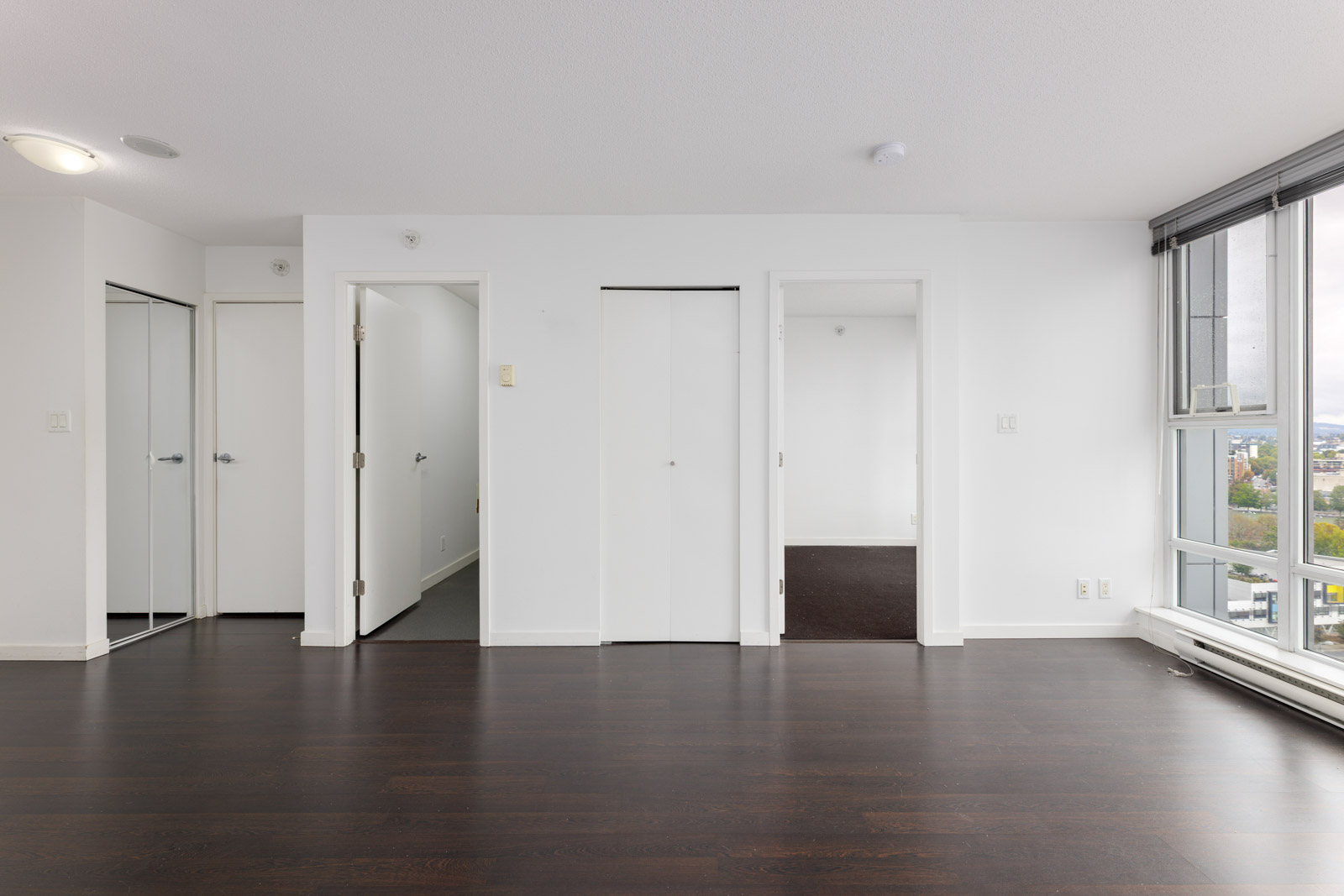 Unfurnished room with dark wood floors, white walls, three doors, mirrored closet, and large window with city view.