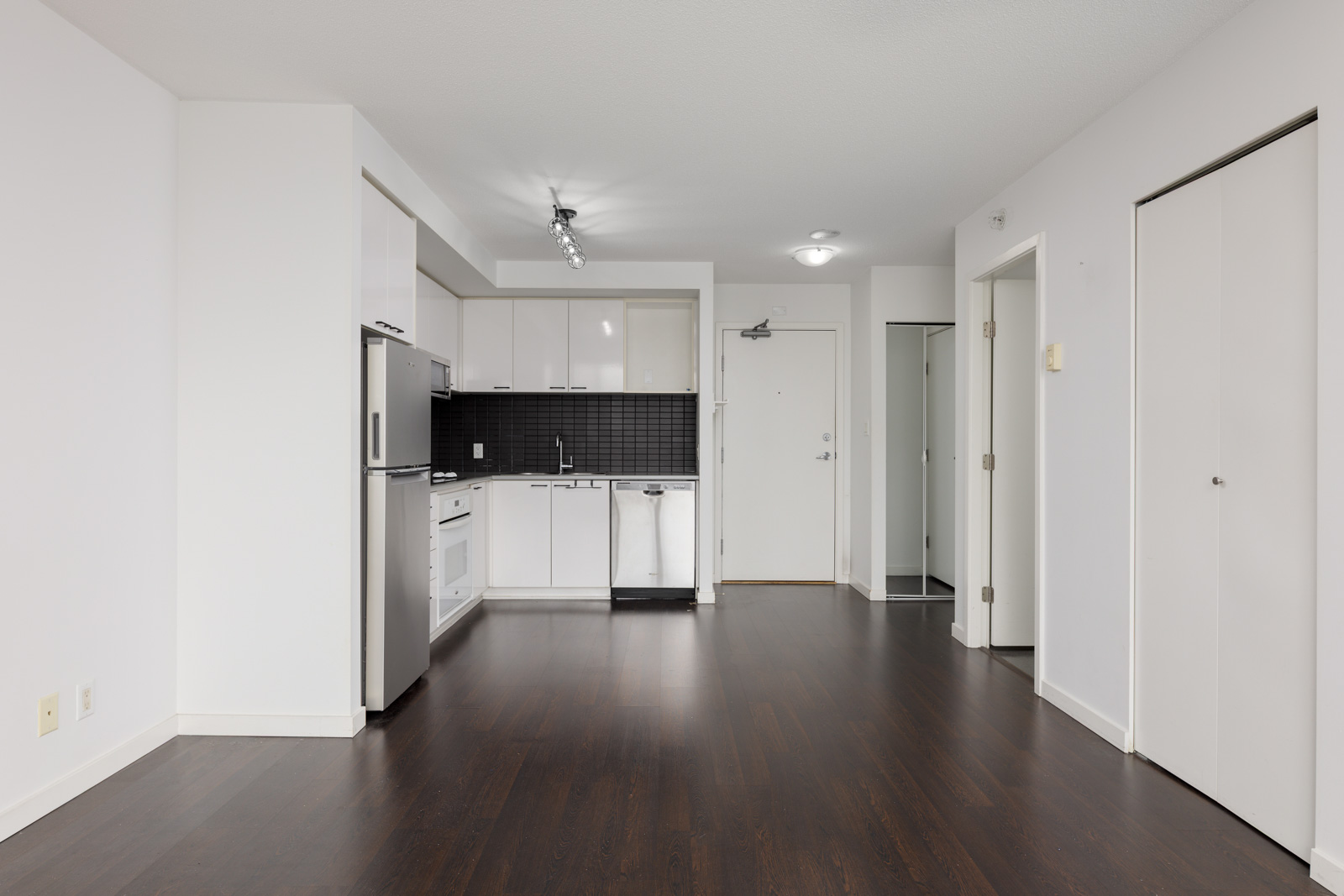 Modern, unfurnished apartment with white walls, dark wood flooring, a compact kitchen area with stainless steel appliances, and multiple doors leading to other rooms.