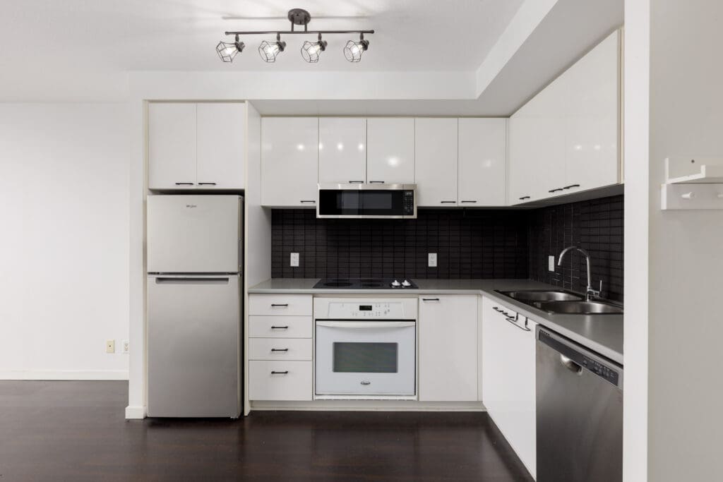 Modern kitchen with white cabinets, stainless steel appliances, black tile backsplash, and dark wood flooring under bright ceiling lights.