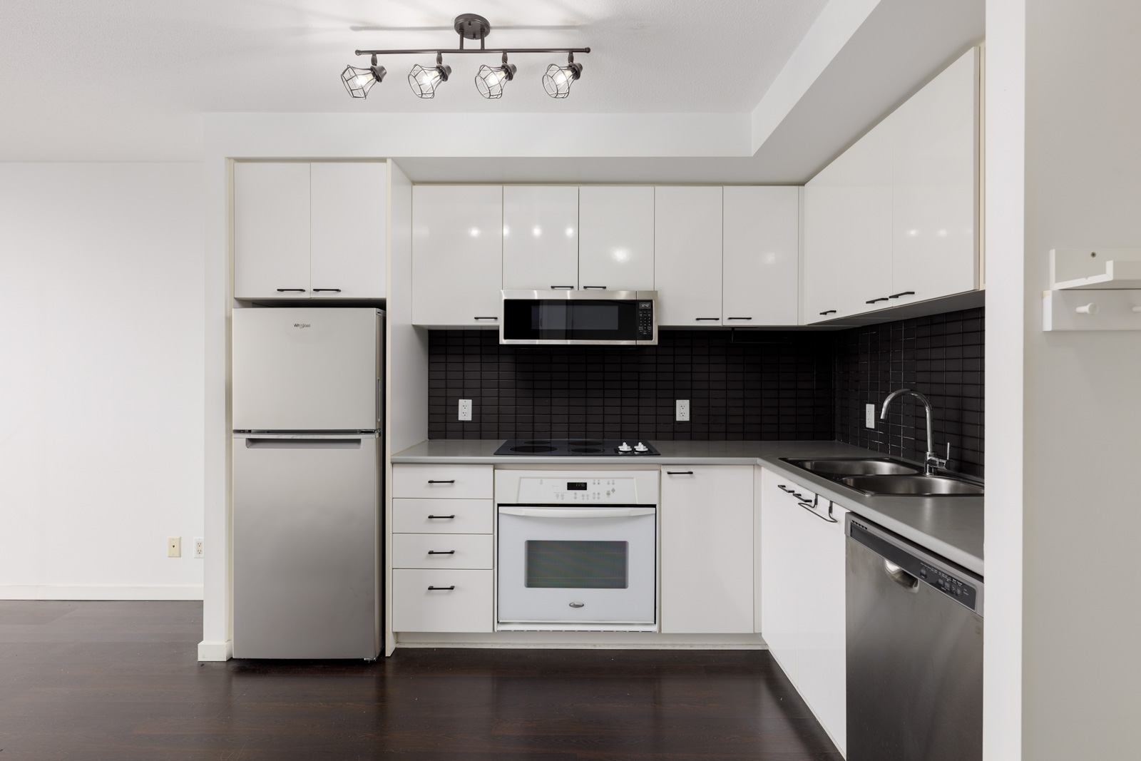 Modern kitchen with white cabinets, stainless steel appliances, black tile backsplash, and dark wood flooring under bright ceiling lights.
