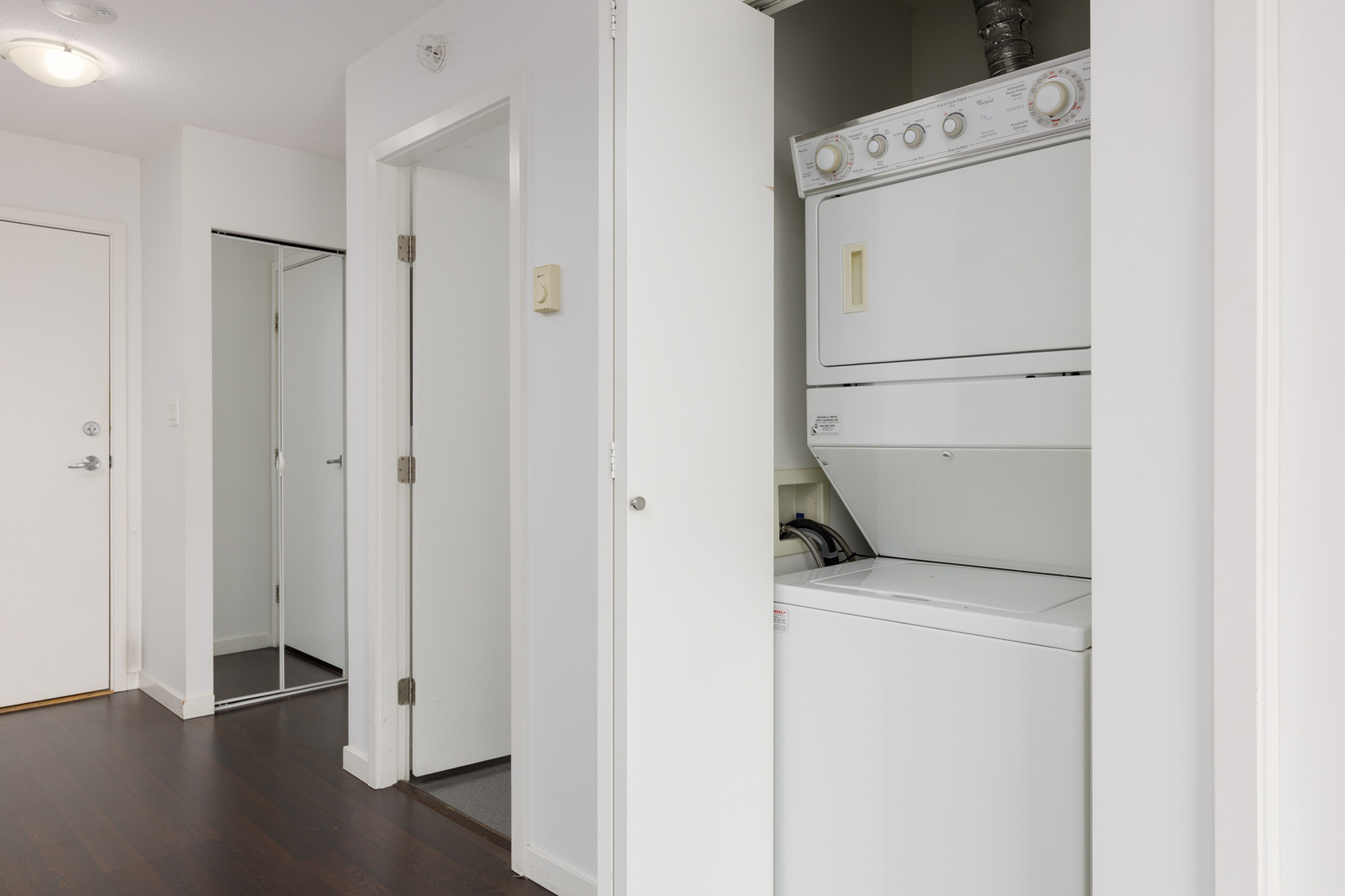 A stacked washer and dryer are installed in a small closet with an open door in a modern apartment hallway with dark wood floors and white walls.