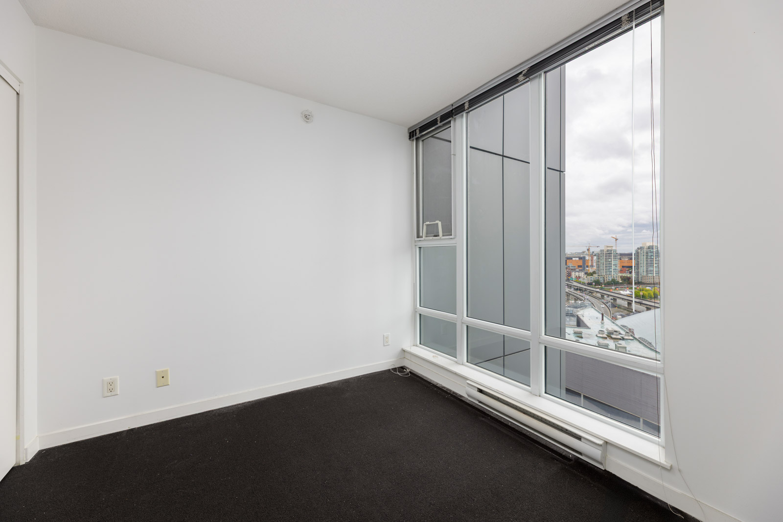 Empty room with white walls, black carpet, and large floor-to-ceiling windows overlooking a cityscape and cloudy sky.