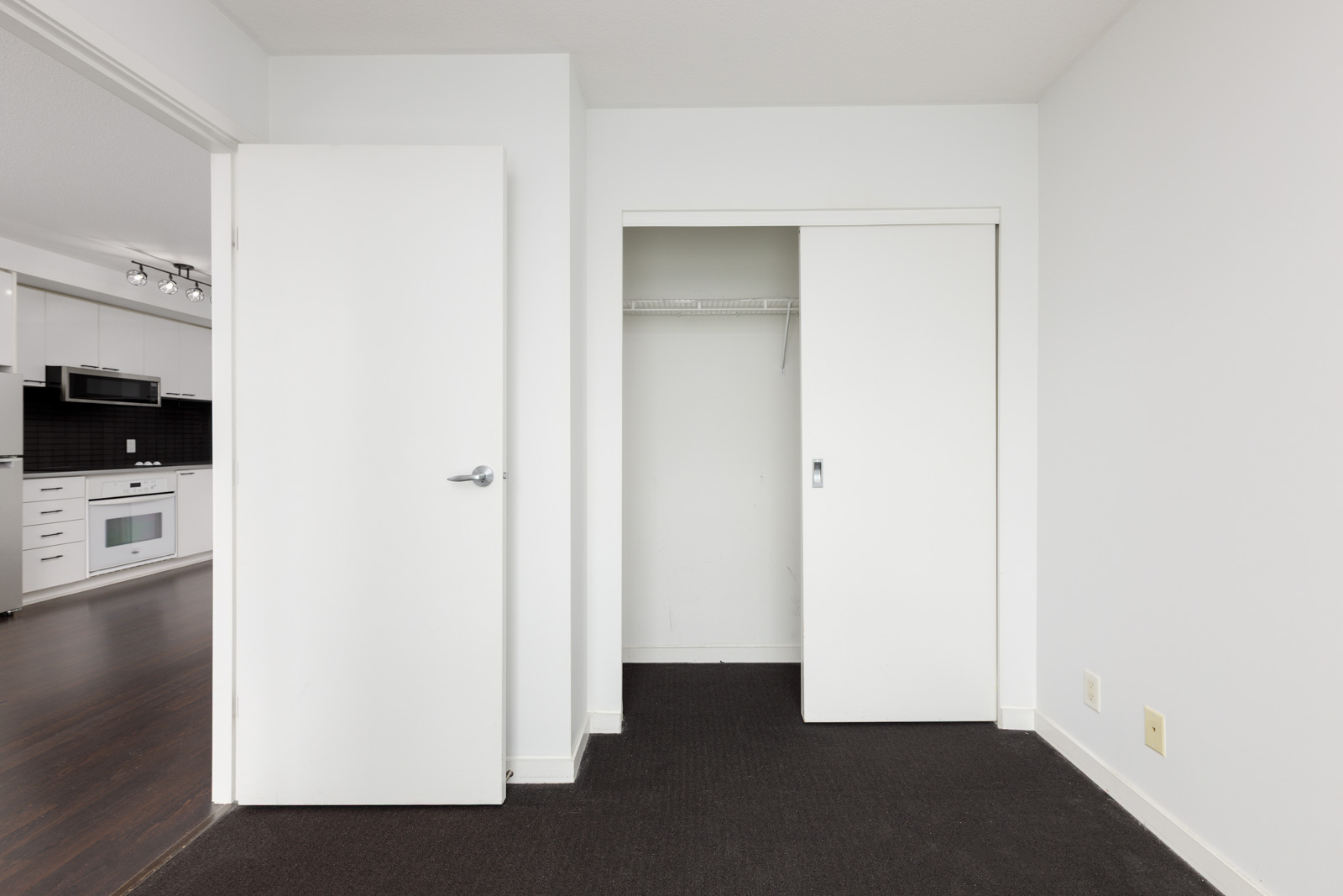 Empty room with white walls, dark carpet, an open closet with sliding doors, and a door leading to a kitchen area with white cabinets and appliances.