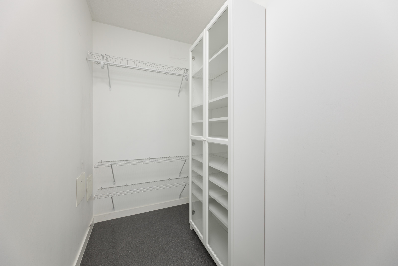Empty walk-in closet with white wire shelves on the left wall and a white storage cabinet with glass doors on the right side.