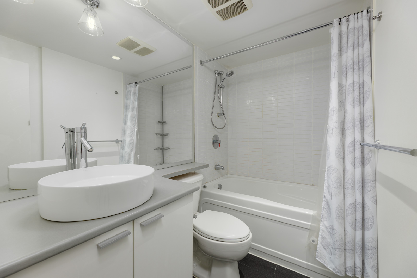 Modern bathroom with a white sink, countertop, toilet, bathtub-shower combination, large mirror, and a white shower curtain with leaf patterns.