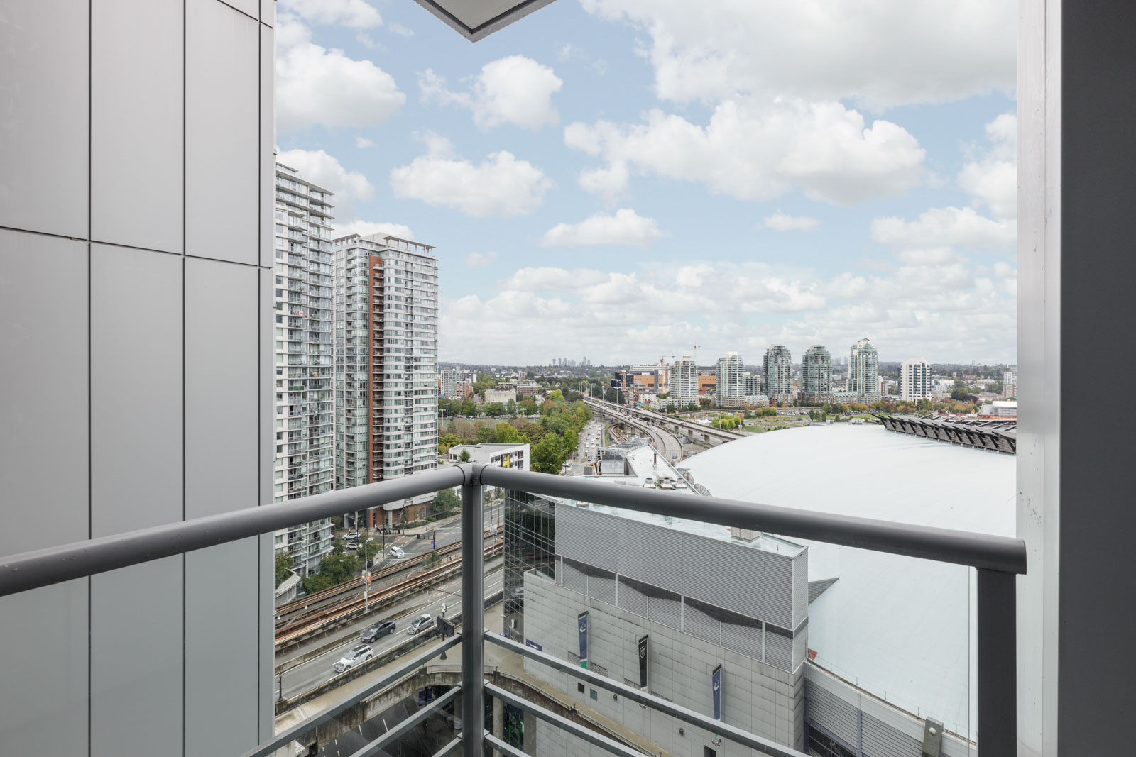 View from a high-rise balcony showing nearby modern buildings, a curved roof structure, roads, railway tracks, and a partly cloudy sky.