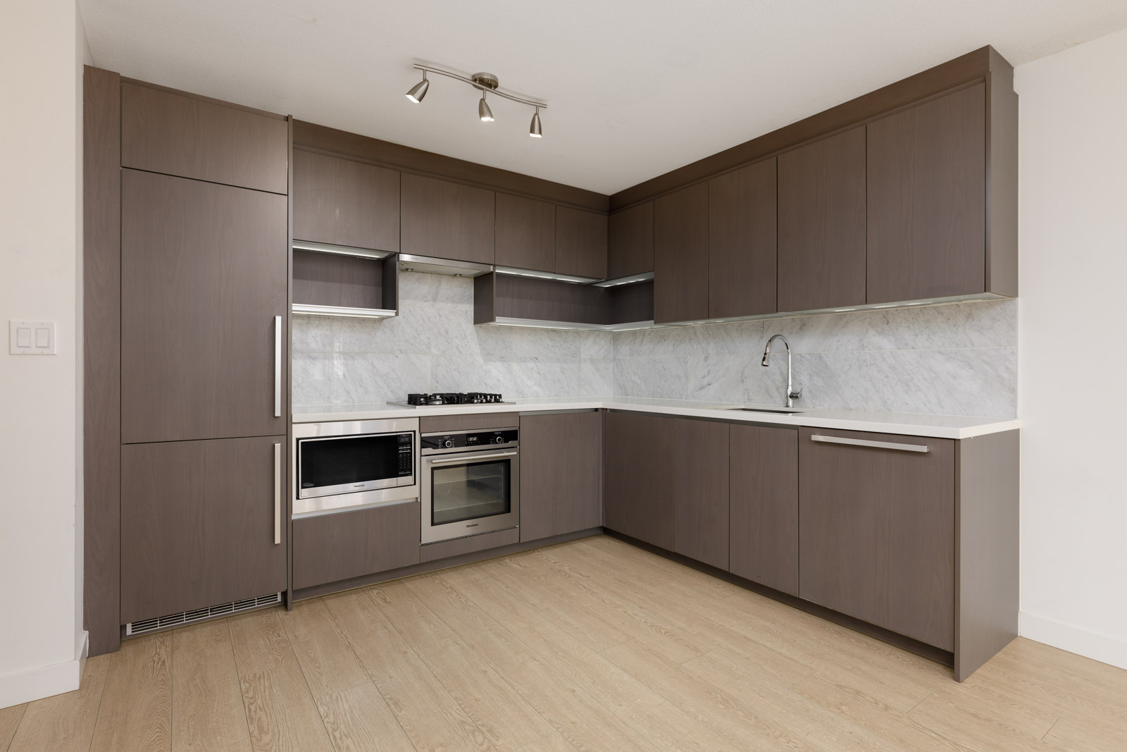 Modern kitchen with brown cabinets, marble backsplash, built-in appliances, and light wood flooring under bright ceiling spotlights.