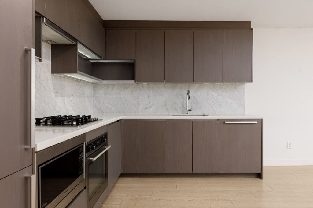 Modern kitchen with brown cabinets, marble backsplash, built-in oven, gas stove, under-cabinet lighting, and a stainless steel sink.