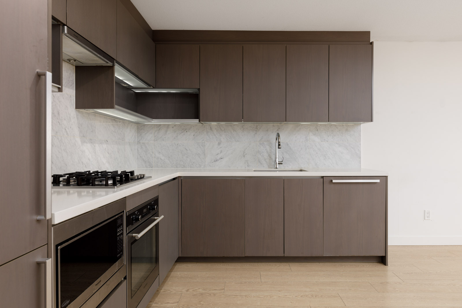 Modern kitchen with brown cabinets, marble backsplash, built-in oven, gas stove, under-cabinet lighting, and a stainless steel sink.