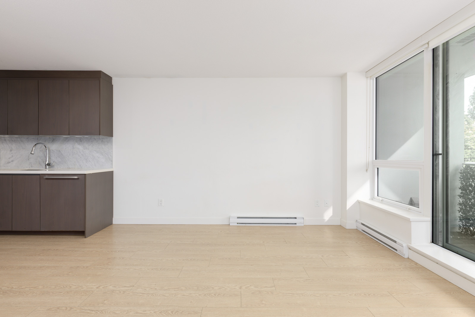 Unfurnished apartment with light wood floors, white walls, a kitchenette with dark cabinets on the left, and large windows on the right letting in natural light.