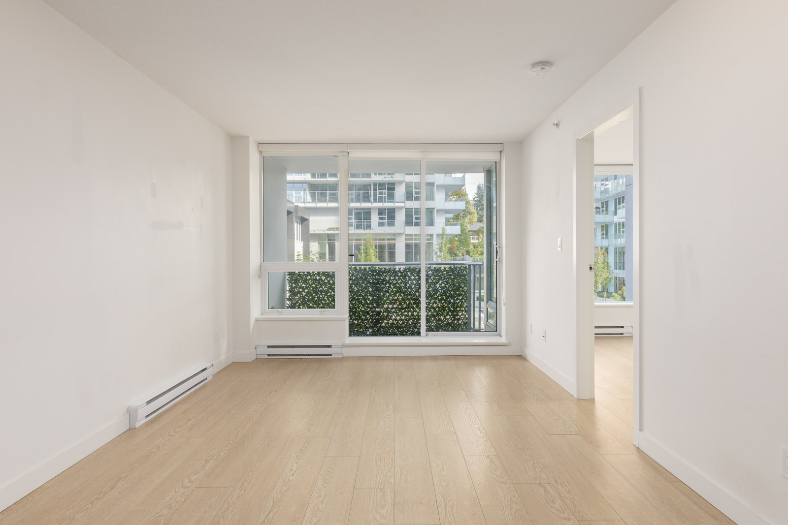 Empty room with light wood flooring, white walls, a large window and glass door leading to a balcony, with an apartment building visible outside.