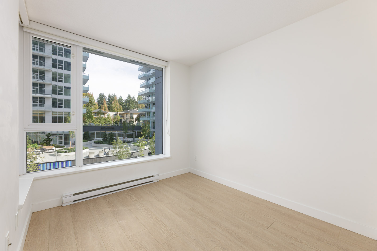 Unfurnished room with light wood flooring, large window, and view of modern apartment buildings and greenery outside.