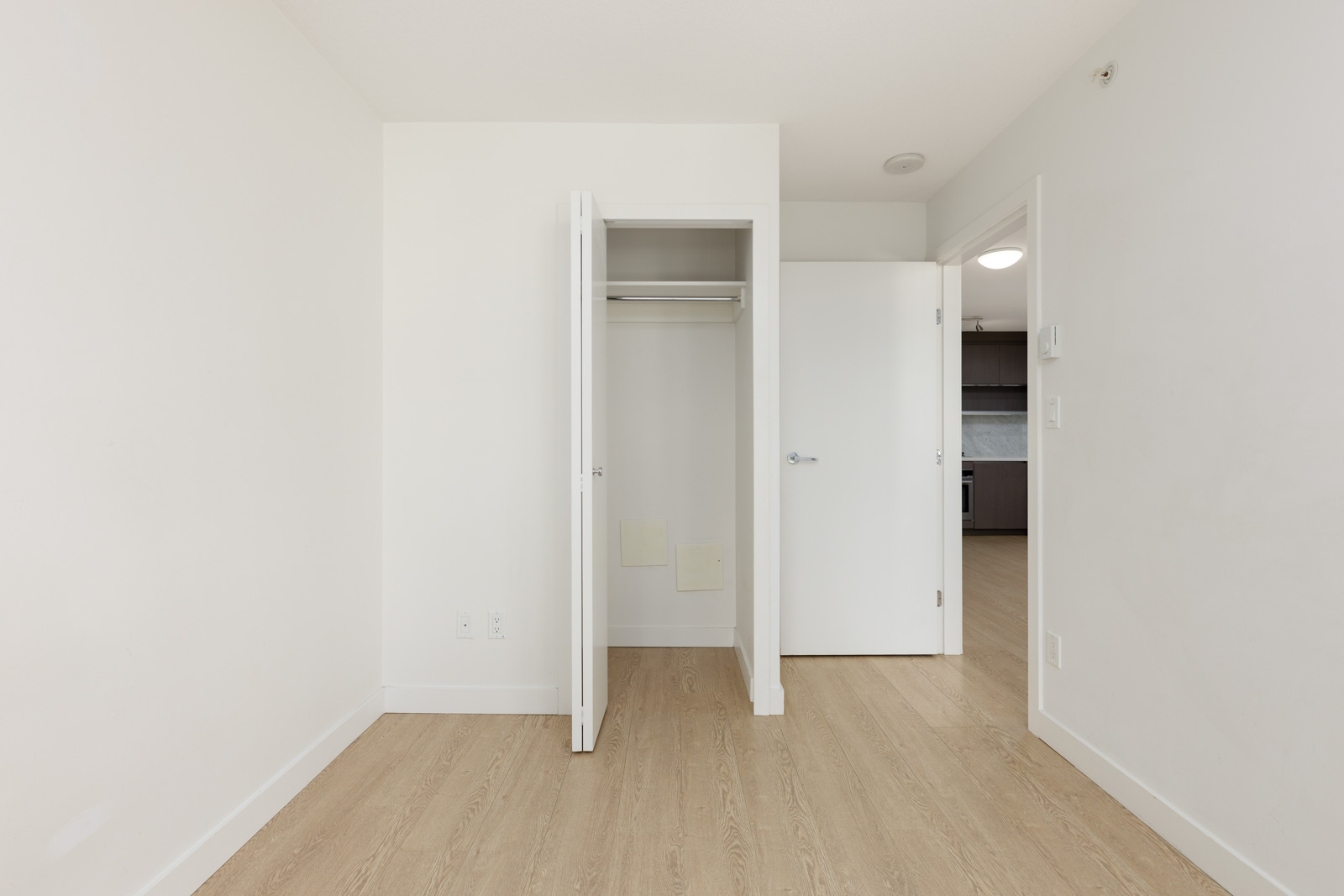 Empty room with light wood flooring, white walls, an open closet with double doors, and an open door leading to a kitchen area.