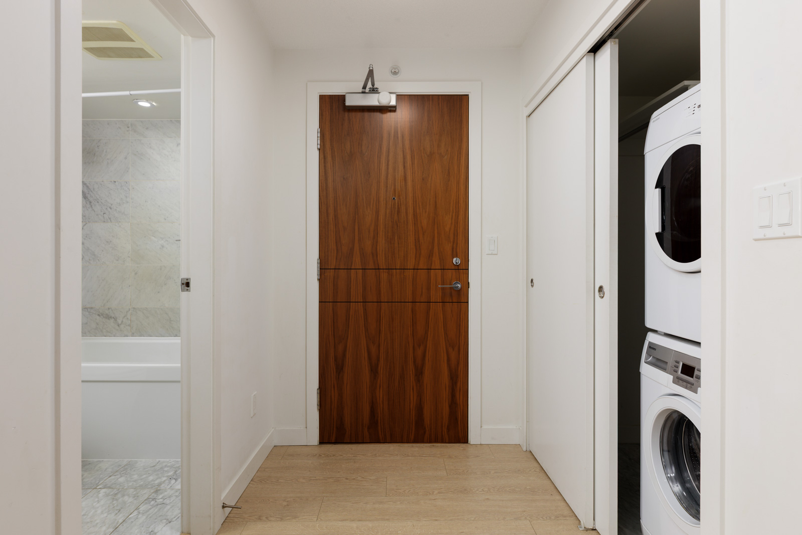 A hallway with a wooden door at the end, a bathroom to the left, and a stacked washer and dryer behind sliding doors to the right.