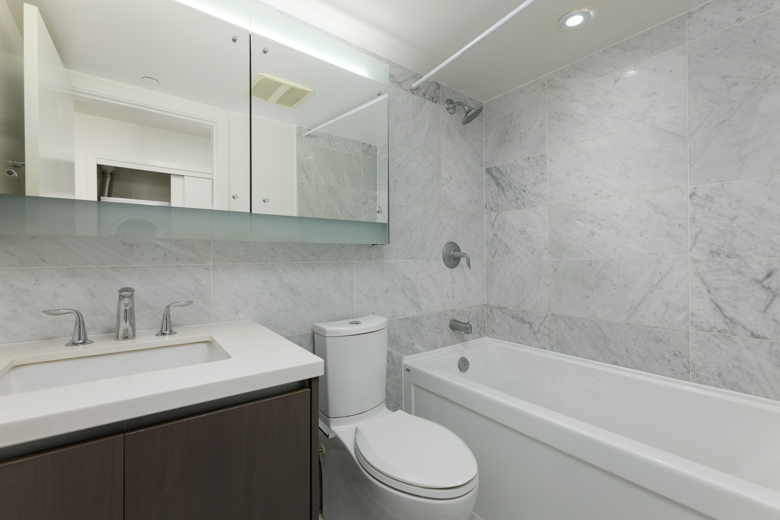 Modern bathroom with white marble tile walls, a built-in bathtub with shower, a toilet, sink with faucet, and mirrored cabinets above the vanity.
