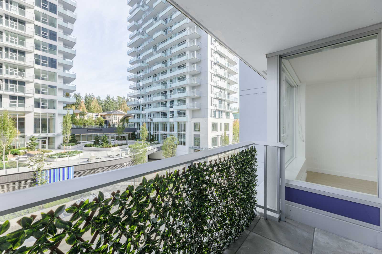 Balcony with green privacy screen overlooks modern apartment buildings and landscaped courtyard on a clear day.