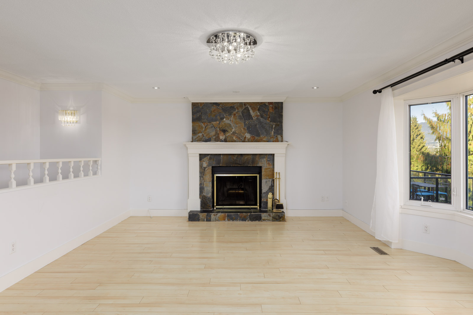 Empty living room with light wood floors, a stone fireplace, white walls, a ceiling light fixture, and large windows with white curtains.