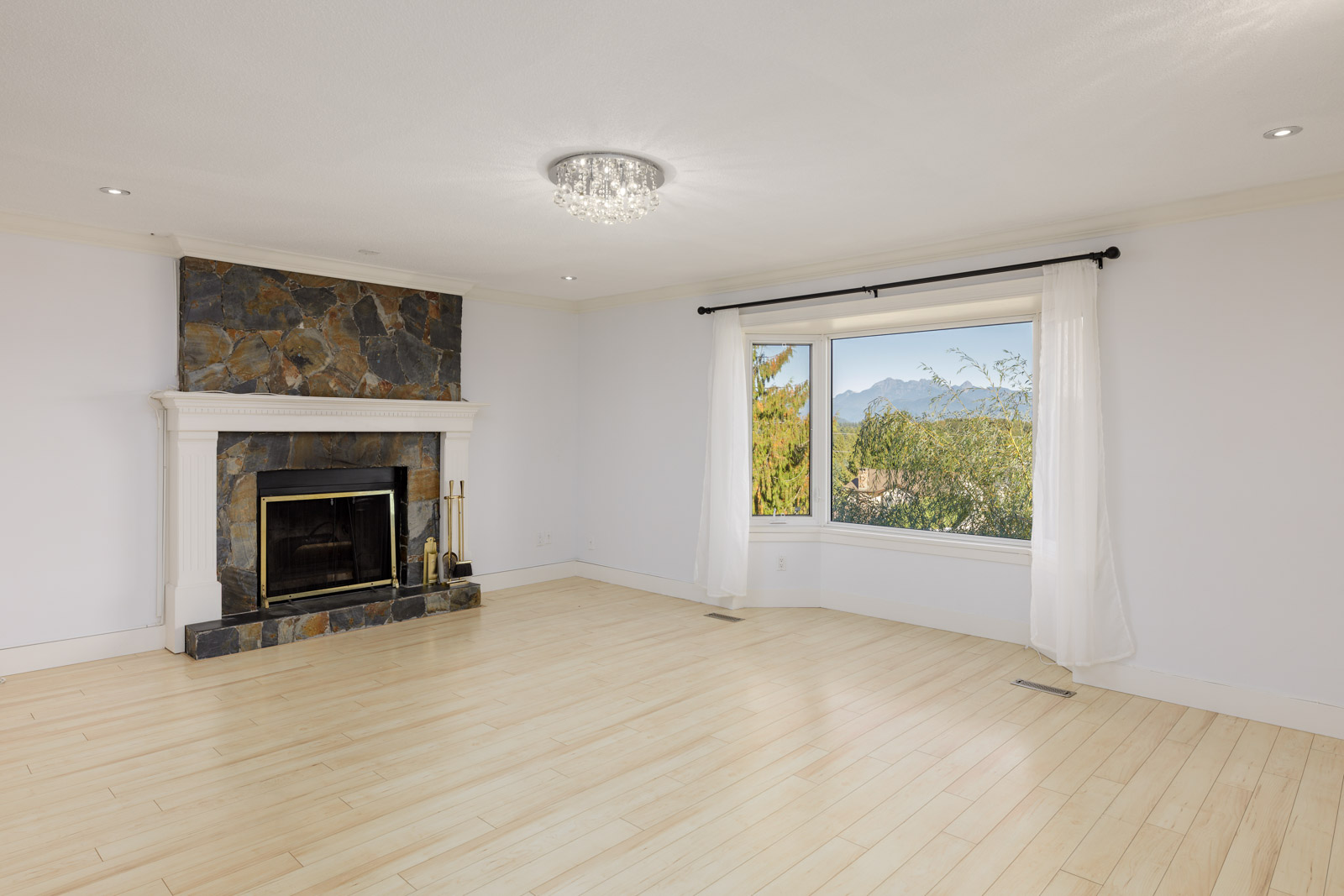 Spacious, empty living room with light wood flooring, a stone fireplace, a large window with white curtains, and mountain views outside.