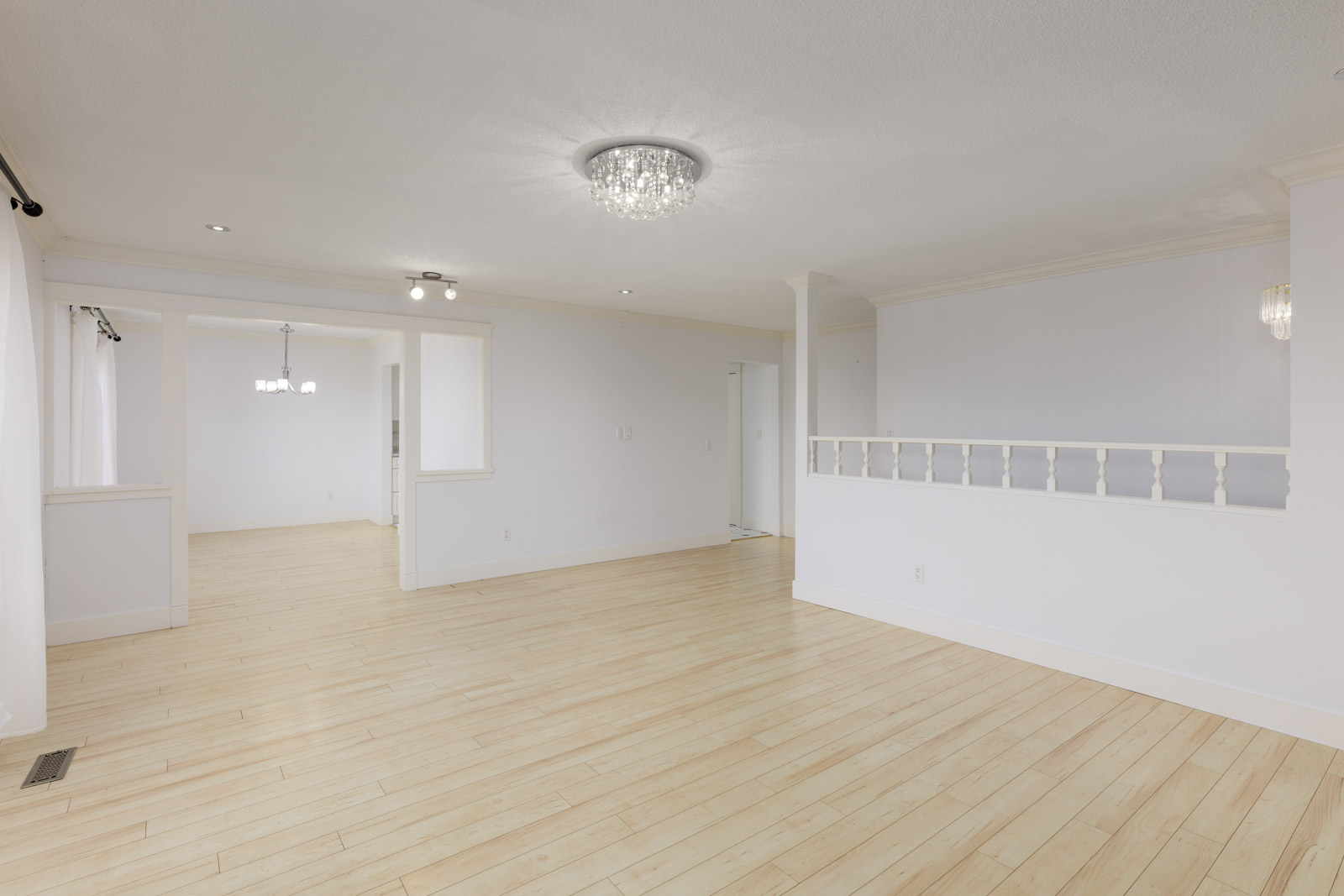 Spacious empty living area with light wood flooring, white walls, ceiling lights, and a partial room divider with railing. An adjacent dining area is visible in the background.