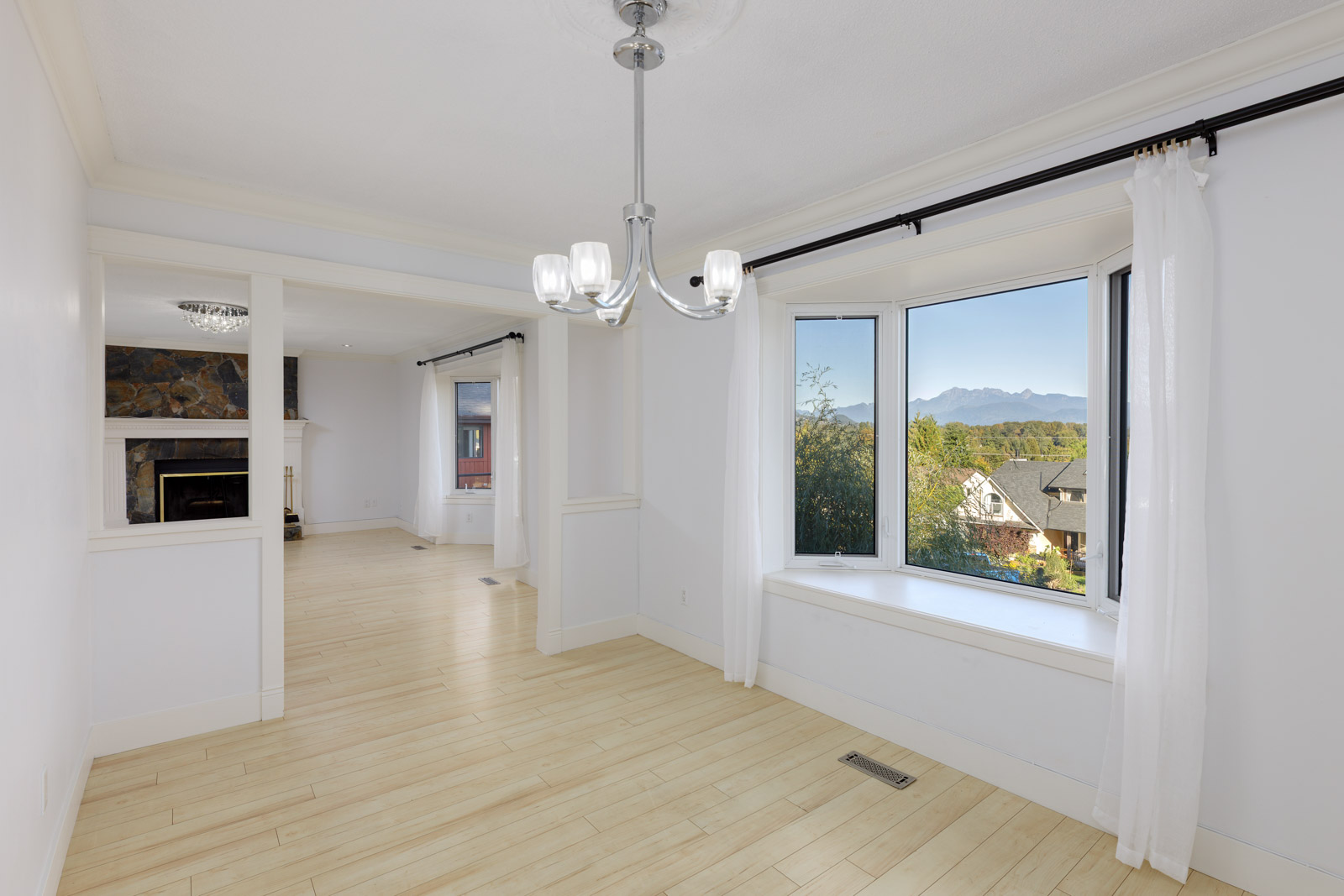 Bright, unfurnished room with light wood floors, large bay window with white curtains, and a view of mountains and trees; adjacent room has a fireplace and stone accent wall.
