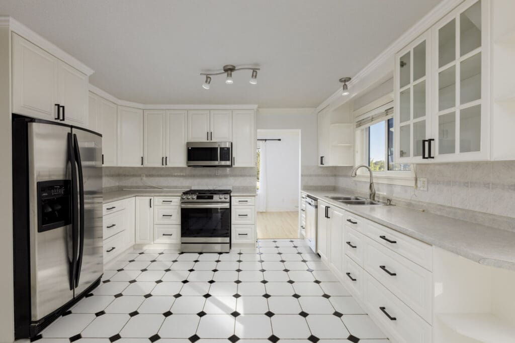 Modern kitchen with white cabinets, stainless steel appliances, black and white tile floor, and under-cabinet lighting. Sink is beneath a window; room is clean and well-lit.