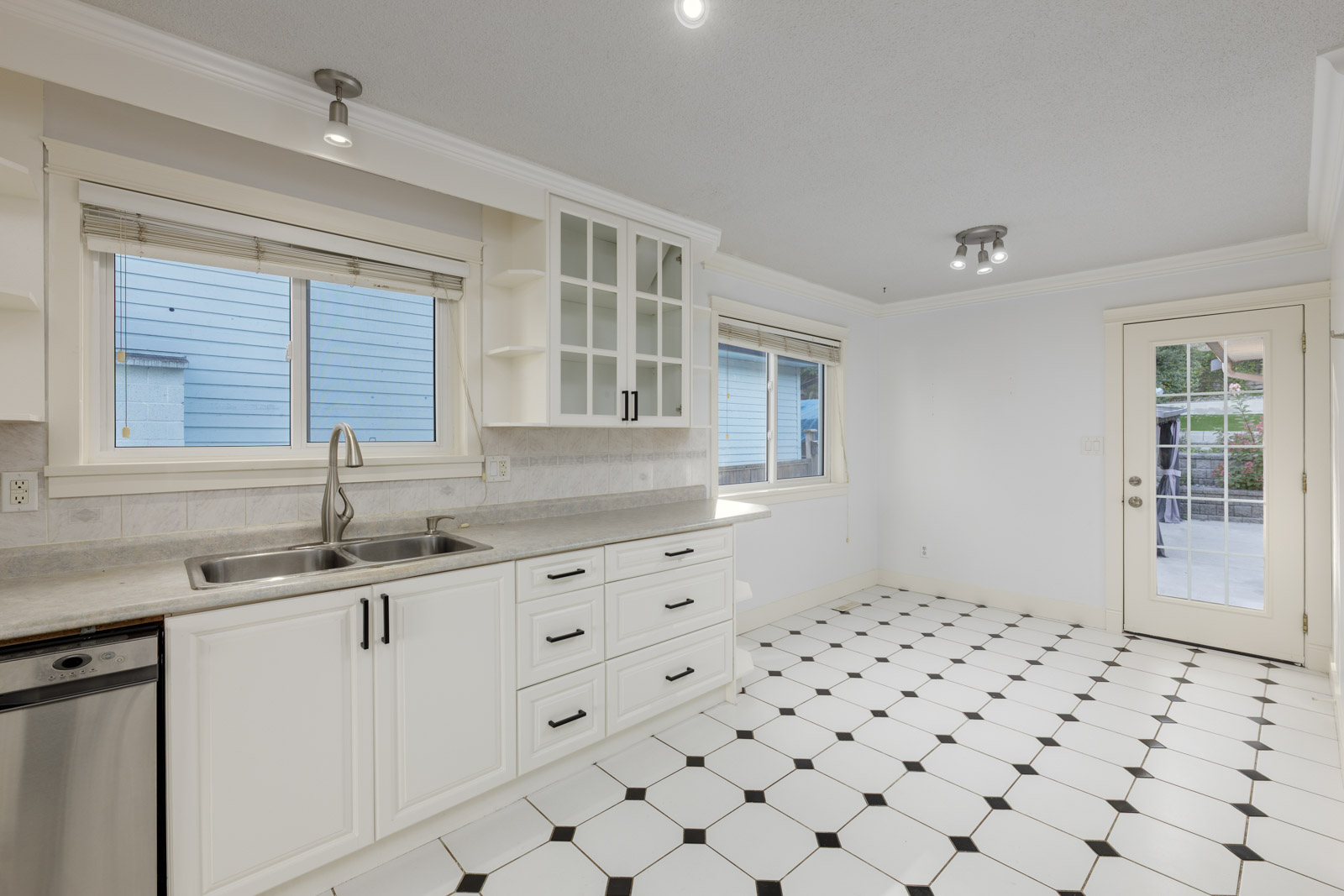 Bright kitchen with white cabinets, black handles, a stainless steel sink, large windows, and a white tile floor with black accents. Door leads to an outdoor patio.