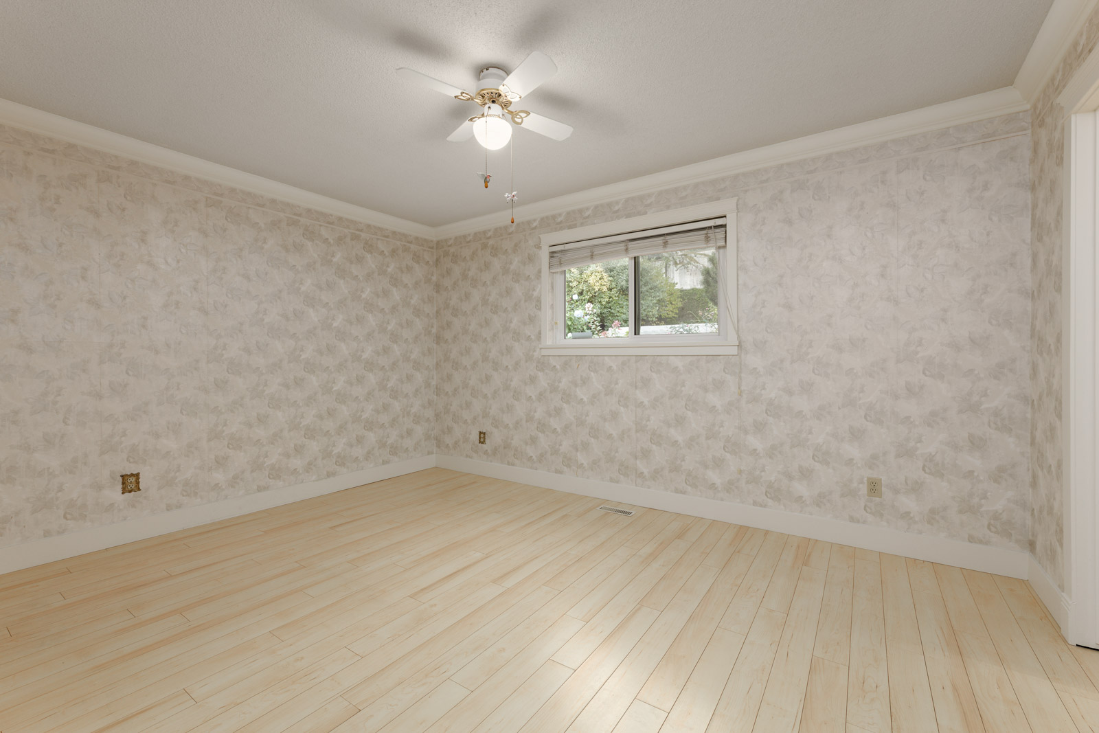 Empty room with light wood flooring, patterned wallpaper, a small window with blinds, and a ceiling fan with a light fixture.