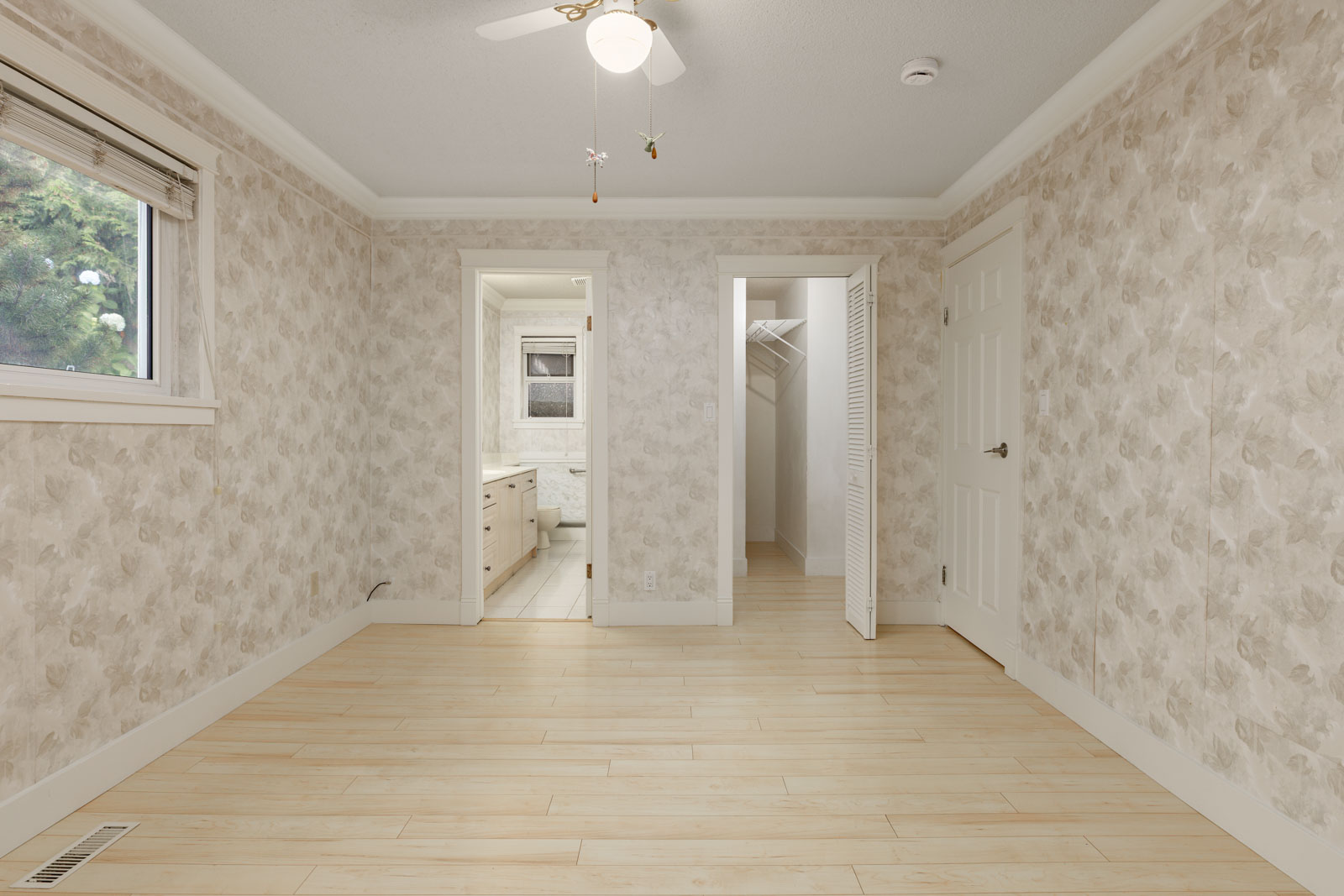Empty room with light wood flooring, patterned wallpaper, a ceiling fan, a window, and doorways leading to a bathroom and a walk-in closet.