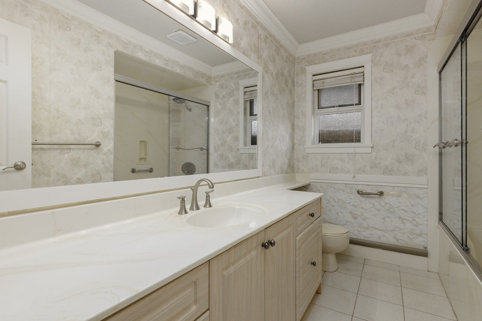 Bathroom with a large mirror, single sink, light wood cabinets, white countertop, toilet, and a glass-enclosed shower; patterned walls and tiled floor.