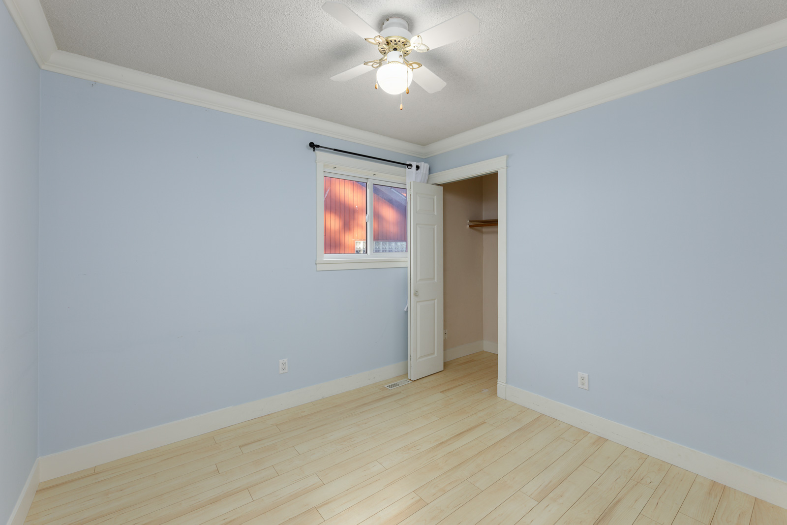 Empty room with light blue walls, beige wood flooring, a ceiling fan with light, a window with blinds, and an open closet door.