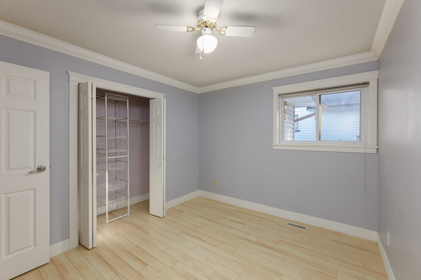 Empty room with light purple walls, light wood flooring, a ceiling fan with light, an open closet with wire shelves, and a window showing a partial outdoor view.