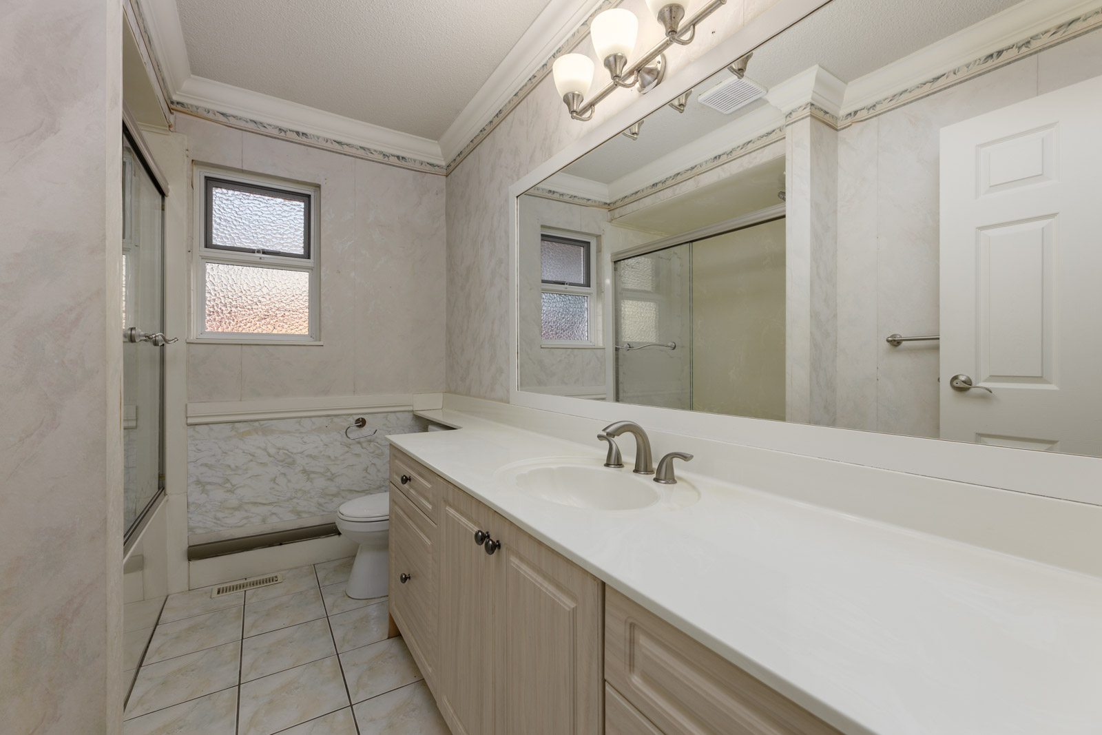 A bathroom with a long countertop, single sink, large mirror, toilet, tiled floor, and a glass-enclosed shower.