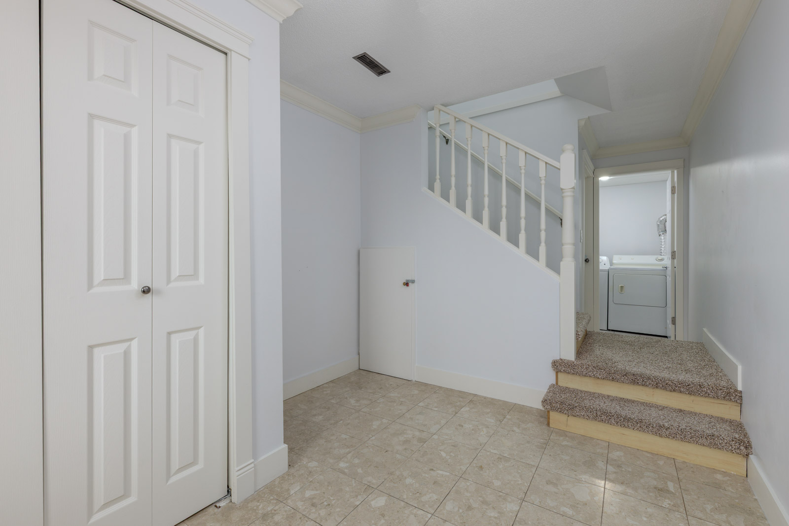 Basement hallway with tile flooring, carpeted stairs, a white closet door, a small storage door, and a laundry area with washer and dryer visible in the background.