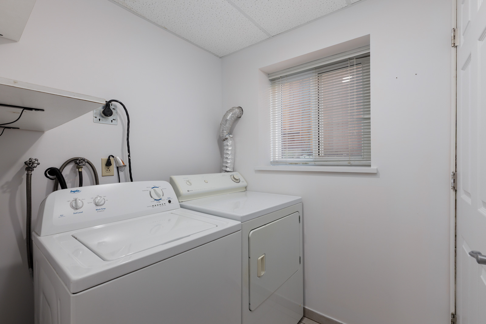 A laundry room with a white washing machine and dryer side by side, a small window with blinds, and white walls and ceiling.