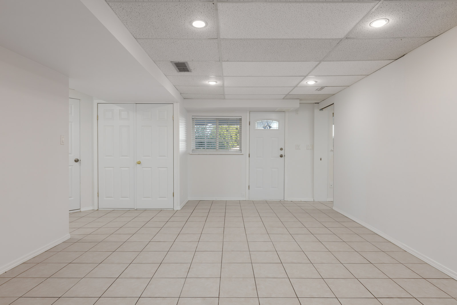 A clean, empty room with tiled floors, white walls, a small window, a door with a window, and white double closet doors. Ceiling lights are recessed into tiled panels.