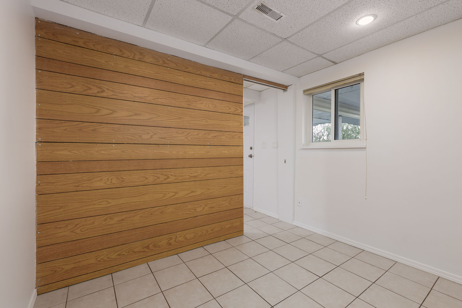 A small room with white walls, tile flooring, a wooden accent wall, a window with a beige shade, and a white door in the background.