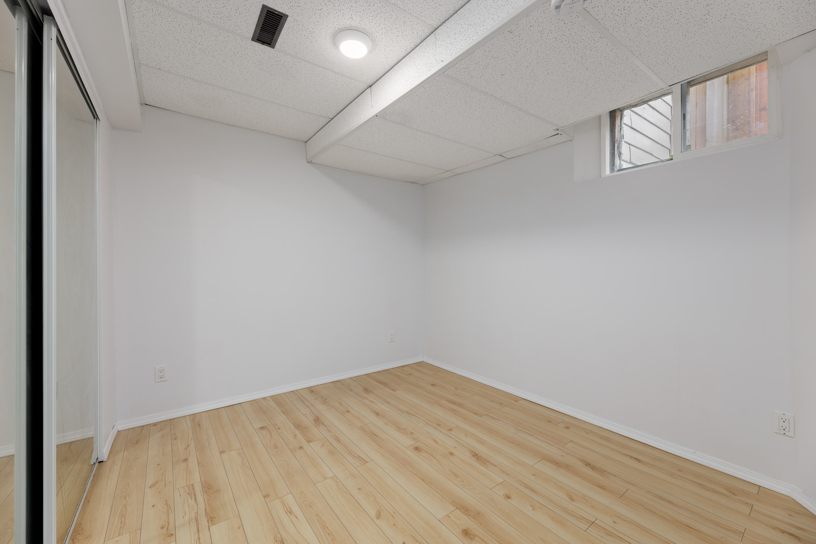 Empty room with light wood flooring, white walls, a small window, and ceiling vents. There are sliding mirrored closet doors visible on the left side.