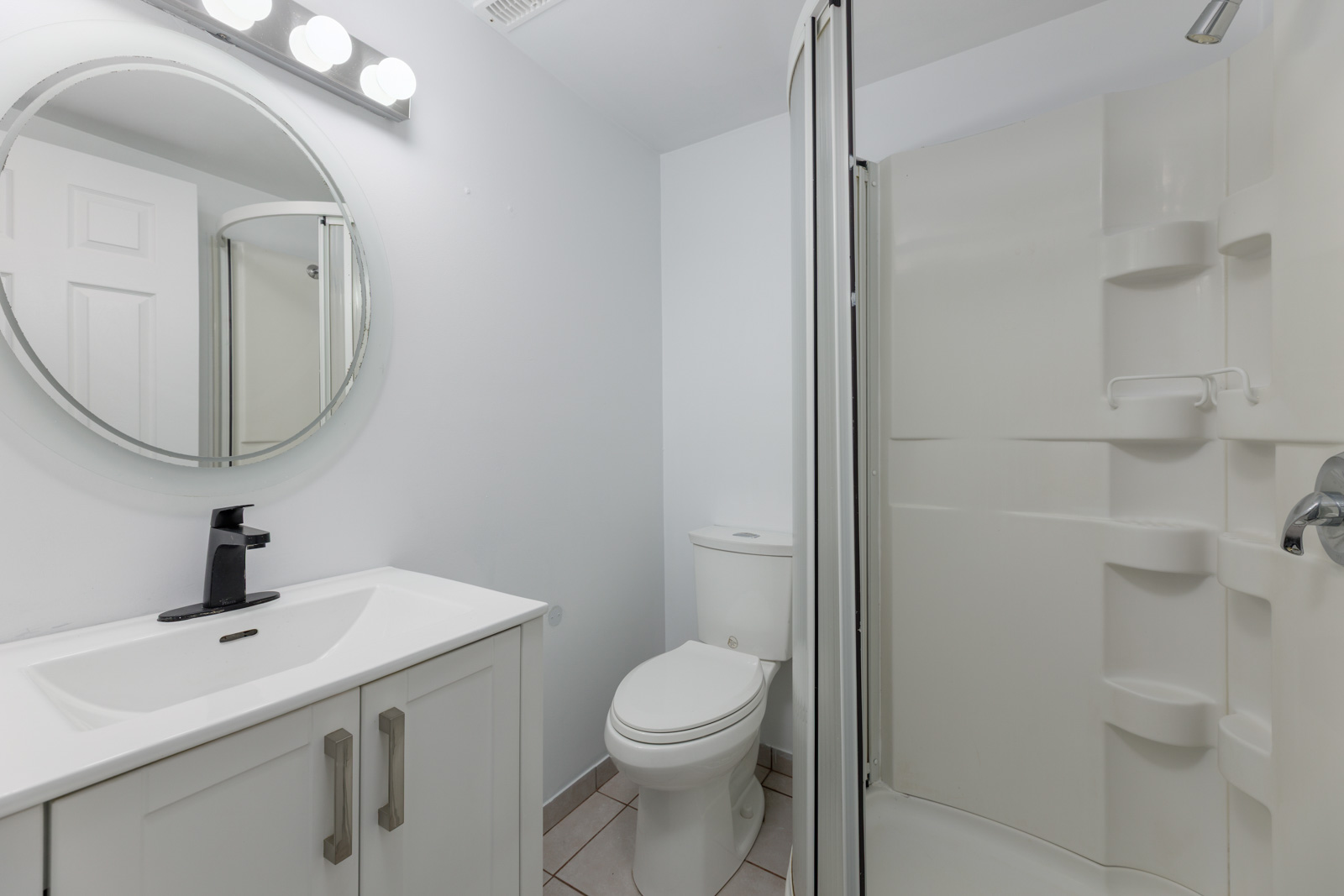 Modern bathroom with a white vanity, round mirror, toilet, and corner shower with built-in shelves.