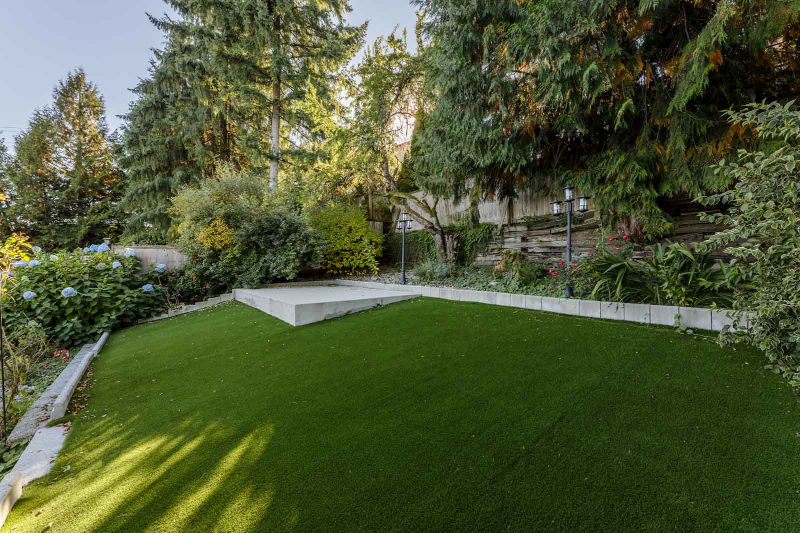 A backyard with a neatly maintained artificial grass lawn, raised concrete patio, dense shrubs, and tall evergreen trees along the fence.
