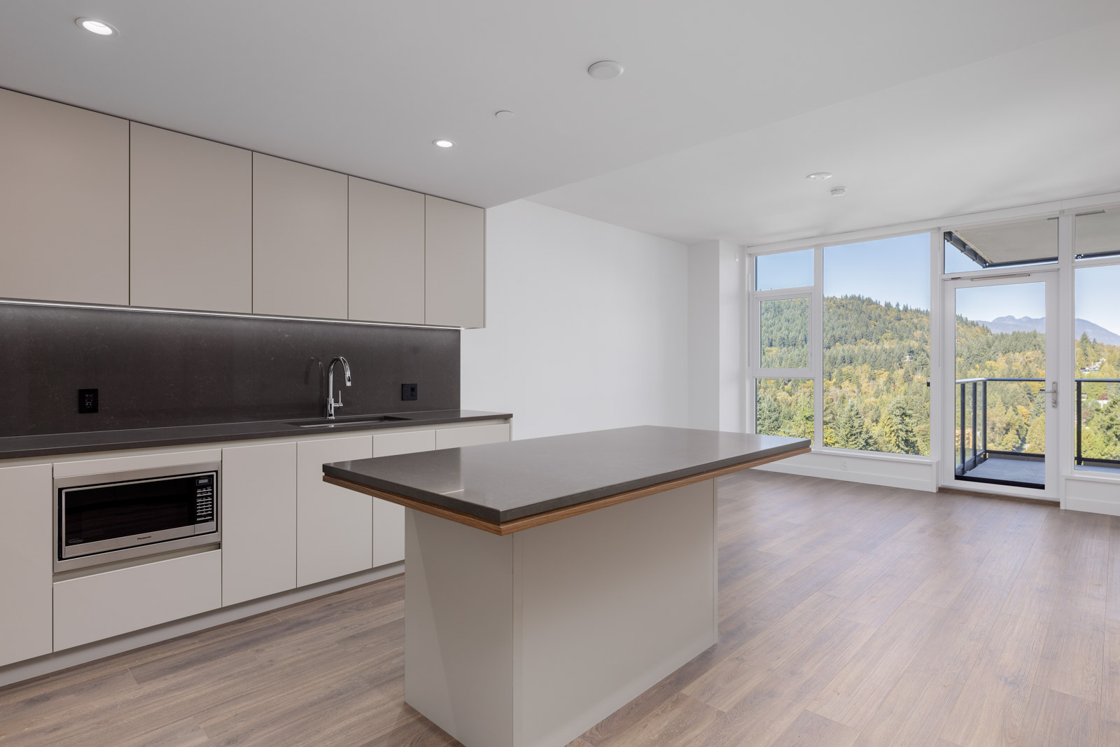 Modern kitchen with beige cabinets, a central island, built-in microwave, and large windows overlooking a green, wooded landscape.