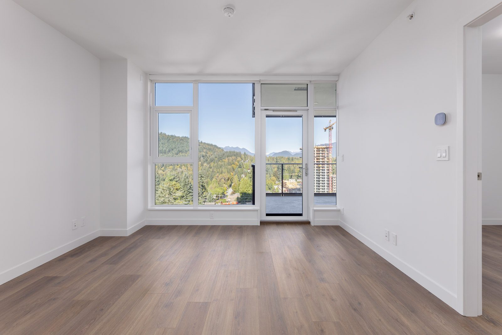Unfurnished room with large windows, glass door to a balcony, white walls, and wood flooring; view of trees, mountains, and buildings outside.