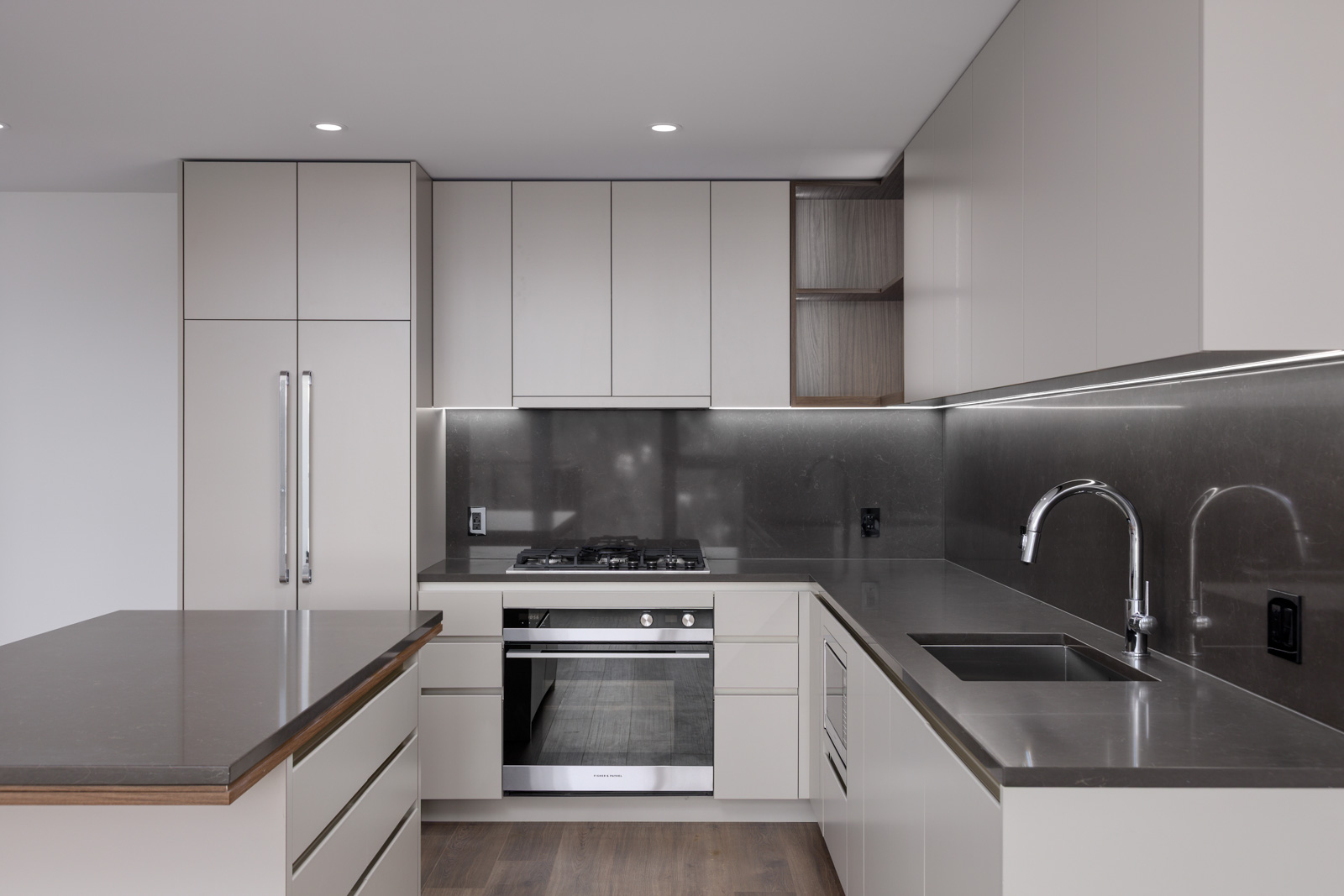 Modern kitchen with light-colored cabinets, stainless steel appliances, built-in oven, gas cooktop, under-cabinet lighting, and a large island with a dark countertop.