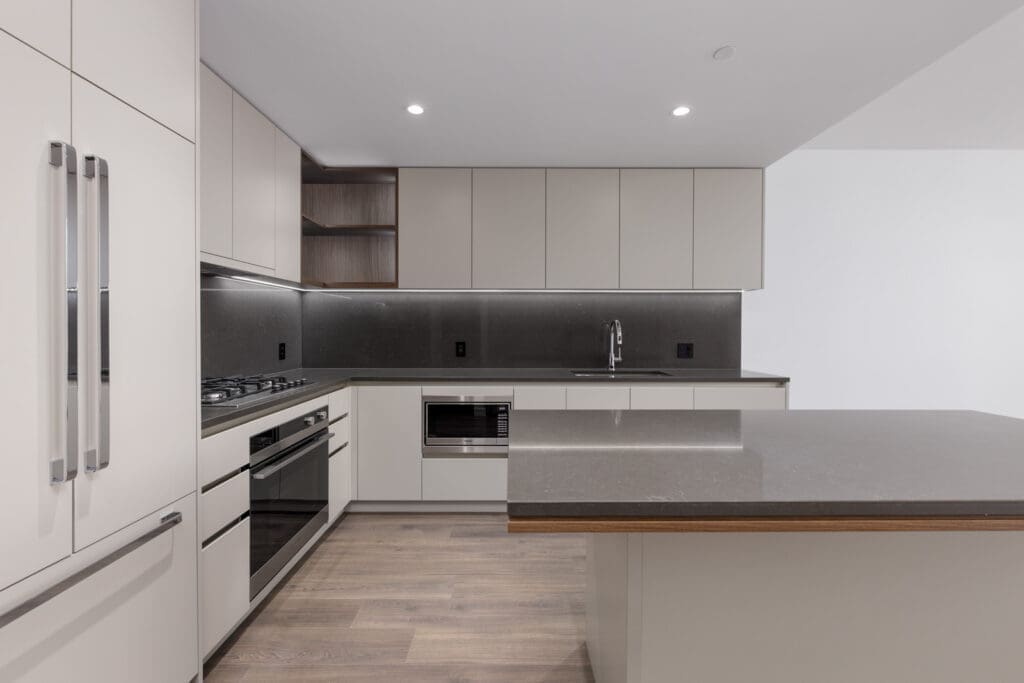 Modern kitchen with light-colored cabinets, dark countertops, built-in appliances, a large island, and wood flooring under bright recessed lighting.