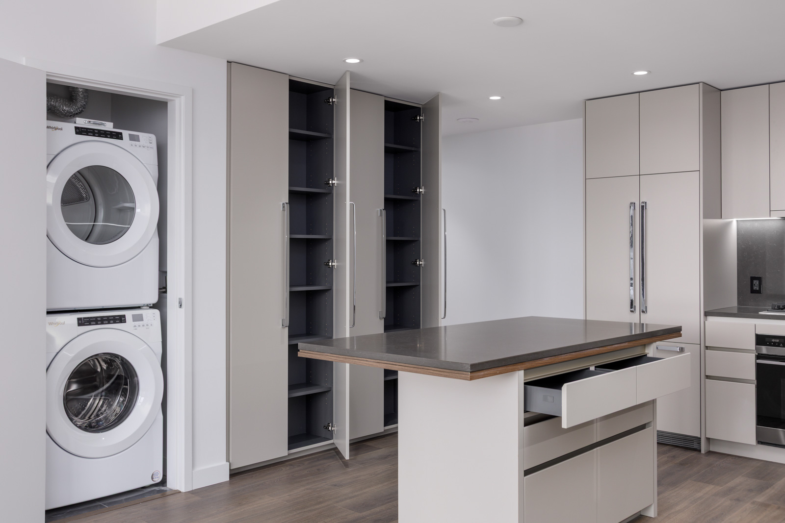 Modern kitchen with light cabinets, a central island, open pantry doors, and a stacked washer and dryer unit in a recessed alcove.
