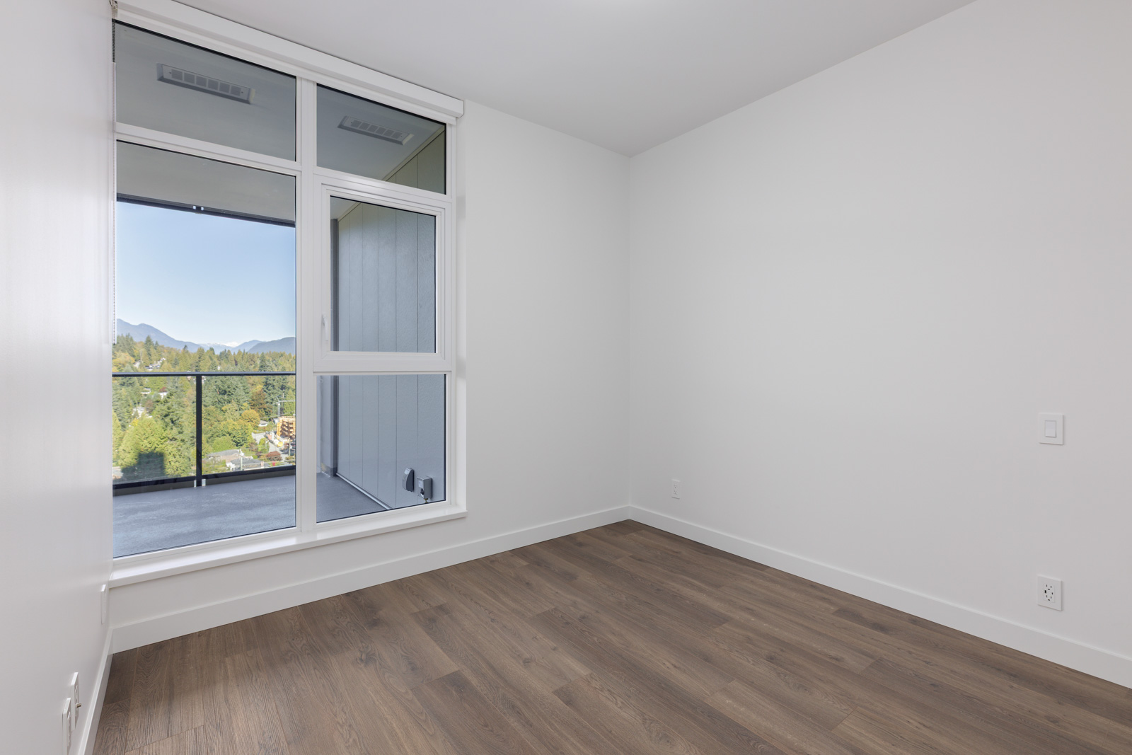Empty room with white walls, wood flooring, and a large window showing a balcony and a view of trees and mountains outside.
