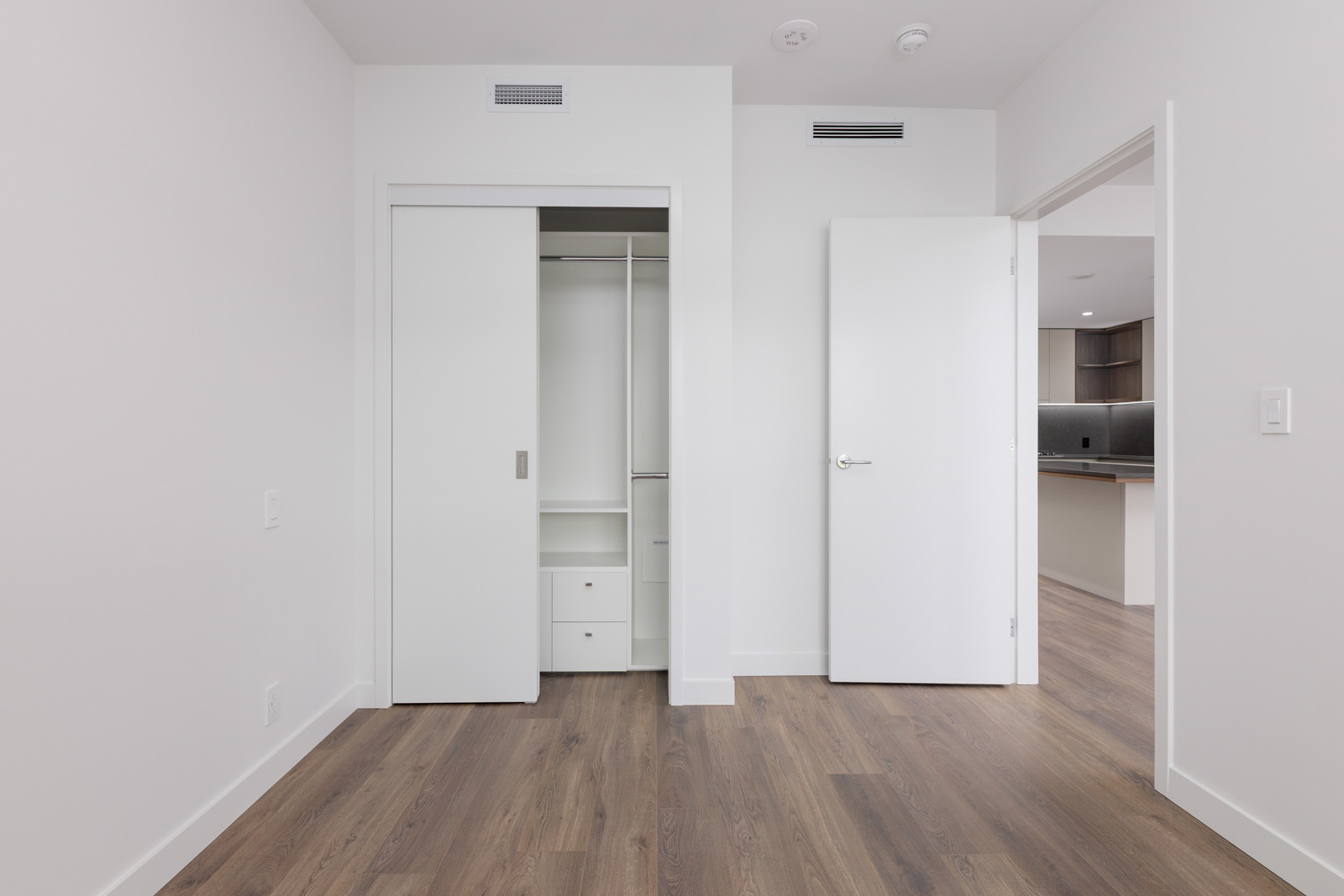 Empty room with white walls, open closet with sliding doors, open door leading to a kitchen, and wood-style flooring.