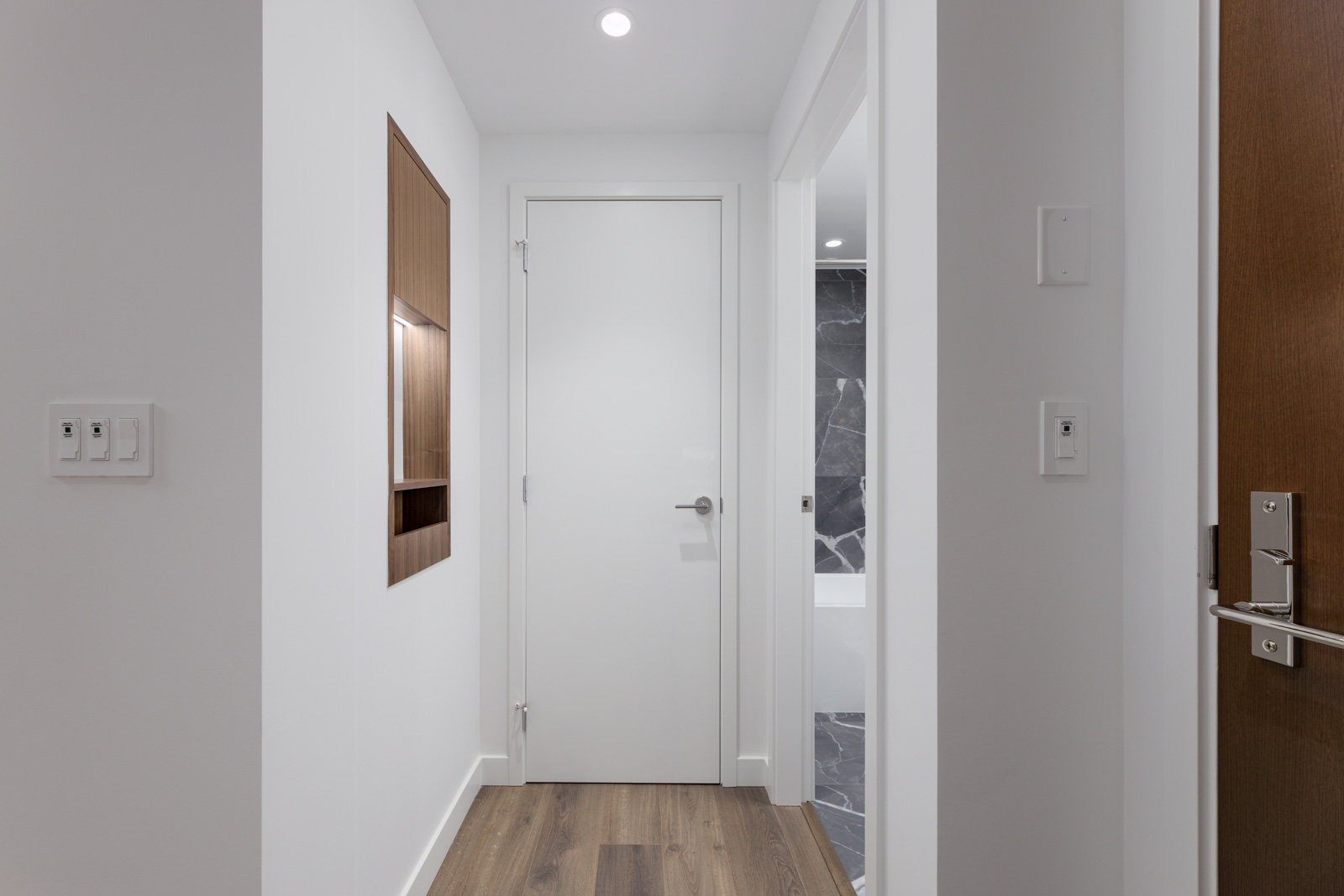 A modern hallway with white walls, a closed white door at the end, wood flooring, and a partial view of a bathroom with dark tile.